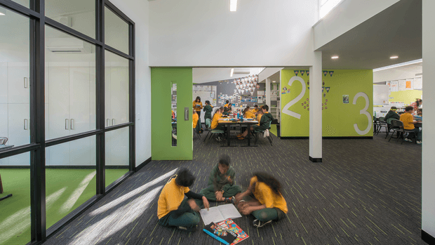 A bright, contemporary open-plan learning or collaborative workspace with lime-green walls marked by oversized numerals, dark carpet flooring, black-framed glazed partitions, and high ceilings with clerestory-style glazing. Students work at tables in the background while a small group collaborates on the floor in the foreground. The space has a playful, flexible educational or commercial interior fit-out.