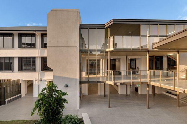 Exterior view of a contemporary multi-storey institutional or commercial building arranged around a paved courtyard. The composition features a glazed central atrium, projecting balconies and walkways with slim metal balustrades, exposed columns, and a contrasting concrete-clad vertical element. The facade combines extensive clear glazing, dark-framed windows, warm beige rendered surfaces, and flat roof forms under a bright blue sky.