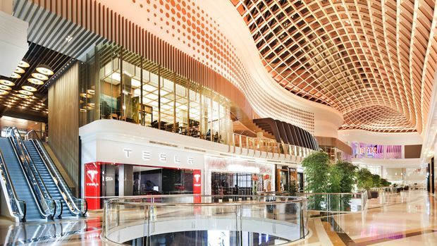 A bright, expansive multi-level commercial shopping mall interior featuring a Tesla retail storefront, escalators, glass balustrades, polished floors, extensive glazing, and a dramatic curved perforated and gridded ceiling installation with integrated lighting.