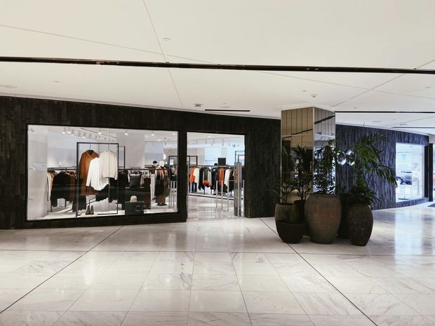 A contemporary retail shopping centre interior with a polished light stone-tile floor, dark textured feature walls, expansive glass shopfronts displaying clothing, mirrored columns, recessed lighting, and large indoor planters. The broad circulation area is bright and open, with a clean, upscale commercial finish.