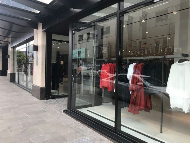 Street-level view of a contemporary retail clothing storefront in a covered shopping arcade. The shop has large black-framed glazed display windows and doors, dark ceiling and wall elements, recessed spot lighting, and clothing racks with predominantly black, red, and white garments. Grey tiled paving extends along the arcade, with reflections of nearby vehicles and buildings visible in the glazing.