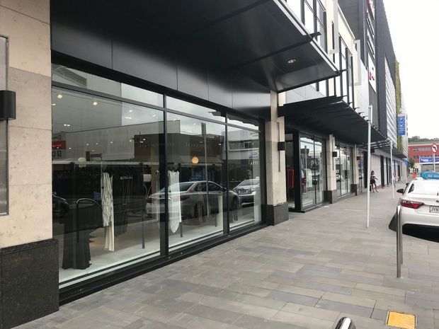 Street-level view of a contemporary commercial building facade with large full-height shopfront glazing, dark metal awnings and cladding, pale stone wall panels, and a wide paved pedestrian sidewalk. Retail displays and reflected street traffic are visible through the windows.