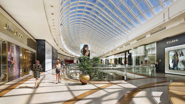 A bright, spacious upscale shopping mall interior with a sweeping curved glass-and-metal barrel-vault skylight, polished tiled floors, glass balustrades, indoor planting, and luxury retail storefronts including Jimmy Choo and Escada. Strong sunlight creates geometric shadows across the concourse.