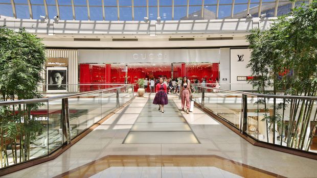 Bright upscale shopping centre atrium with a glazed roof, polished stone flooring, glass-and-metal balustrades, indoor trees, and a pedestrian bridge leading toward luxury retail storefronts including Gucci and Louis Vuitton.