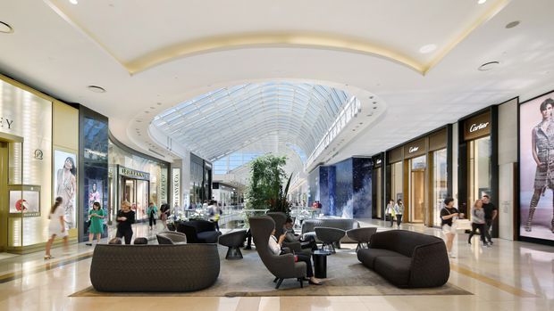 A bright, upscale multi-level shopping mall interior with luxury retail storefronts, polished stone flooring, sculptural lounge seating, indoor planting, and a large curved glass skylight forming a dramatic central atrium. The space has a refined contemporary commercial design with abundant daylight and spacious circulation.