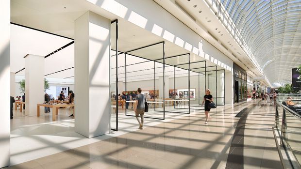 A bright, expansive contemporary commercial interior resembling a shopping centre or gallery, with a long glazed barrel-vaulted roof, extensive skylighting, white structural columns, polished pale flooring, and large black-framed glass retail or display enclosures. People and shopfronts line the broad circulation corridor, with strong geometric shadows cast across the floor.