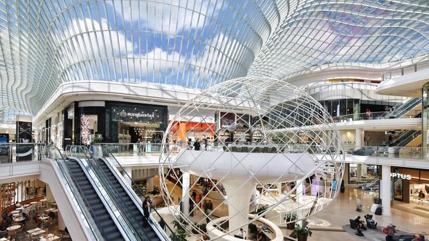 A bright, expansive multi-level shopping centre atrium with retail stores, escalators, open balconies, and a dramatic curved glazed roof. A large white sculptural architectural feature with a geometric metal lattice occupies the centre of the space, creating a contemporary commercial interior.