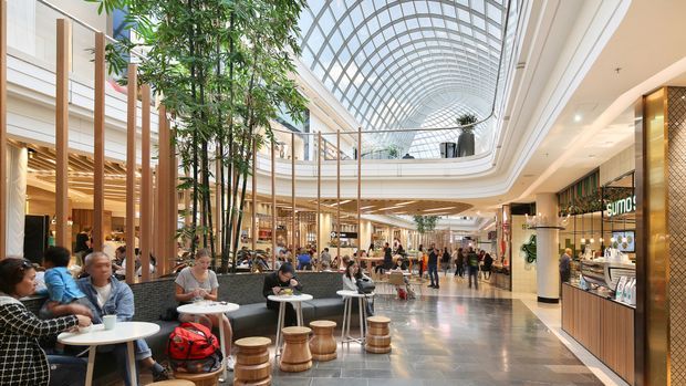 A bright, spacious multi-level commercial shopping and dining atrium with a dramatic curved glazed roof, upper-level balconies, extensive timber columns and ceiling detailing, polished stone flooring, retail storefronts, café seating, and abundant indoor planting. The scene is busy and contemporary, with a large open circulation space and a strong indoor-outdoor atrium feel.