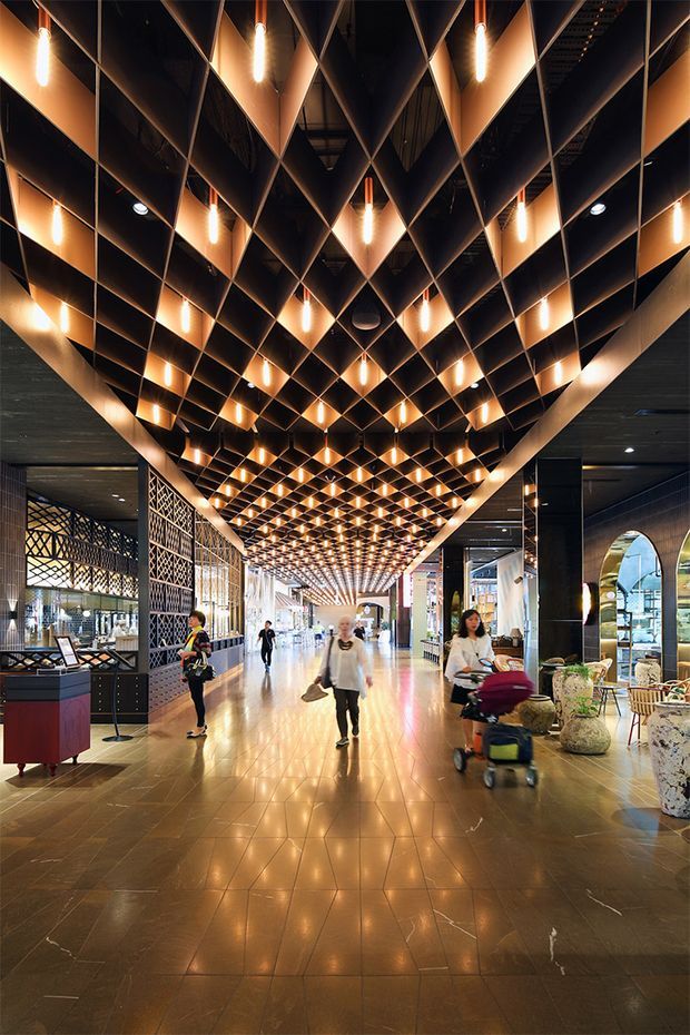 A wide commercial interior corridor or concourse with a dramatic geometric suspended ceiling composed of repeating dark and warm metallic diamond-shaped panels. Numerous warm pendant tube lights are integrated throughout the ceiling, creating a strong rhythmic pattern and glowing amber atmosphere. The polished tiled floor reflects the lighting. Retail or hospitality storefronts, decorative screens, display counters, seating, and several people occupy the sides and distance of the space.