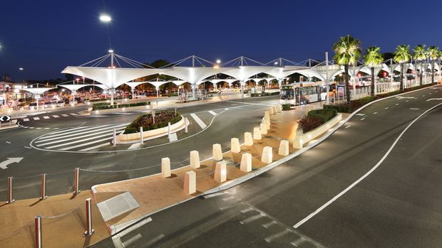 Wide nighttime exterior view of a modern commercial transport or shopping precinct, featuring sweeping white tensile canopy structures, illuminated pedestrian areas, landscaped islands, palm trees, road markings, and multiple vehicle lanes.