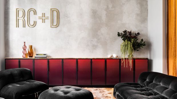 A stylish living room with a long, low burgundy-red built-in sideboard against a textured grey plaster wall. Black tufted lounge chairs and a matching sofa surround a low round ottoman, while sculptural vases, books, greenery, and a metallic wall logo provide decorative accents. Warm timber-framed glazing or drapery is visible at the right edge.