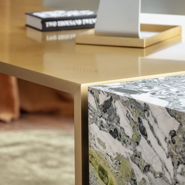 Close-up interior detail of a refined side table or console featuring a warm brass-gold metal frame and a polished, highly veined natural stone surface in white, grey, black, and green tones. A matching stone block or adjoining table element is visible in the foreground, with a book and sculptural decor placed on top. The background is softly blurred and appears to be a sophisticated living area.