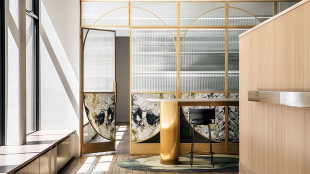 A bright, refined interior featuring a tall glazed screen with warm timber or brass-toned framing, translucent ribbed glass, curved decorative arches, and marble-patterned panels. A sculptural cylindrical column, compact desk or bar-height workspace, pale timber cabinetry, and integrated bench seating create a contemporary architectural fit-out with strong vertical and geometric detailing.