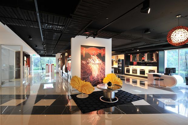 A spacious, highly polished commercial showroom or interior design gallery with a dramatic black ceiling featuring exposed services and track lighting. The reflective geometric tiled floor, dark and cream finishes, built-in display cabinetry, large windows, vivid artwork, sculptural yellow lounge chairs, and a spherical pendant create a bold, theatrical contemporary setting.