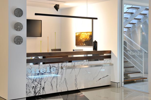 A refined contemporary interior featuring a long marble-faced built-in island or reception-style counter with dark timber open shelving and a white stone surface. A slim linear pendant light hangs above, with wall-mounted artwork and television beyond. Through the glazed partition at right, a modern staircase with white metal balustrades and timber treads is visible. The space uses a restrained white, black, grey, and dark timber palette with polished finishes.