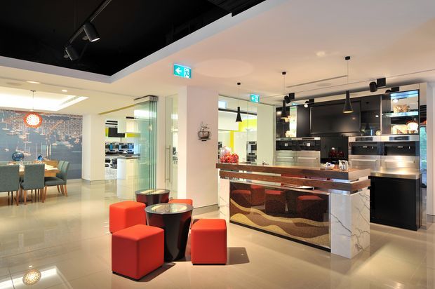 A bright, open-plan commercial showroom interior featuring a prominent contemporary kitchen display with dark cabinetry, integrated appliances, a stone island, and a patterned curved-panel breakfast bar. In the foreground is a casual seating area with red upholstered cube stools and small round tables. A dining display, illuminated feature wall, glass partitions, pendant lights, and black ceiling elements are also visible.