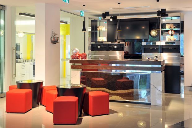A polished contemporary commercial hospitality or entertainment space with a central bar and service counter, glossy dark cabinetry, integrated appliances, display shelving, and a large wall-mounted screen. Bright red cube stools and lounge seats cluster around small dark tables in the foreground. Reflective stone or polished flooring, glass panels, illuminated shelving, pendant lights, and stainless-steel details create a high-contrast, luxurious interior.
