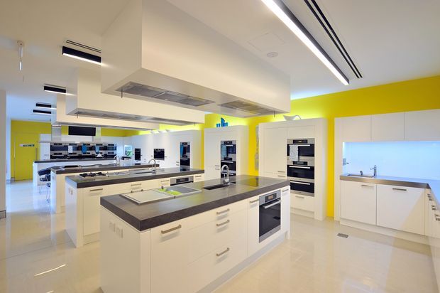Bright, spacious contemporary kitchen showroom or demonstration kitchen with multiple white island workstations, integrated ovens and appliances, dark stone-look benchtops, and extensive white cabinetry. A vivid yellow feature wall and cool blue-lit splashback provide bold colour accents. The ceiling has exposed linear ventilation and large stainless-steel rangehoods, while glossy pale flooring reflects the abundant lighting.