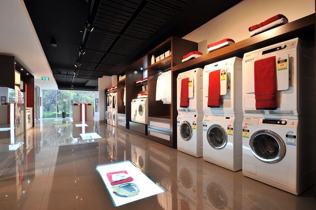 A polished modern retail showroom displaying front-loading washing machines and dryers in rows along dark wood shelving. The space has glossy reflective flooring, bright product lighting, a black exposed-style ceiling with track lighting, and large glazed openings at the rear. Folded towels and laundry products are arranged on the appliances and shelving.