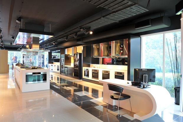 A sleek contemporary kitchen showroom or commercial kitchen display space with multiple built-in kitchen installations, islands, ovens, cabinetry, and benchtops. The space features glossy pale flooring, a black-and-white tiled display zone, dark exposed services and ductwork across the ceiling, strong linear lighting, large windows, and a curved white reception or workstation counter with a computer and bar stool.