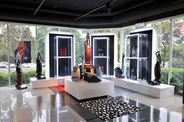 A bright, modern commercial showroom with floor-to-ceiling glazing, glossy tiled flooring, illuminated black display cabinets, white built-in plinths and a central white display island. Vacuum cleaners and related products are arranged throughout the space, with a black-and-white patterned rug and views of landscaped surroundings outside. The ceiling has exposed dark services and track lighting, creating a sleek retail presentation.