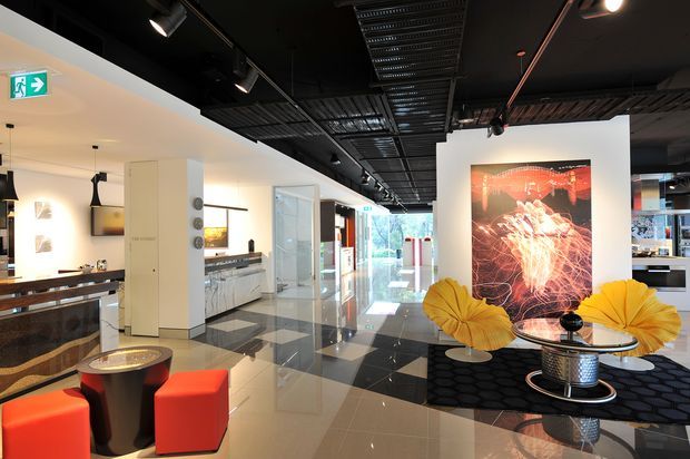 A contemporary commercial interior with a spacious polished floor, black exposed services ceiling, track lighting, and a bright white fit-out. The foreground contains a small lounge arrangement with red cube seating, round tables, and large yellow floral decor, while a reception or bar-like counter and display cabinetry extend along the left side. A large graphic artwork and glazed entry or corridor are visible toward the rear.