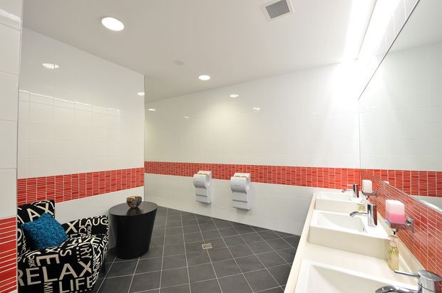A bright, contemporary public or commercial bathroom with multiple white wall-mounted toilets, twin white vanity basins, large white wall tiles, and a distinctive horizontal red mosaic tile stripe. The floor is dark grey tiled, and a small seating area with patterned upholstered chairs and a black cylindrical table occupies the left side. Recessed ceiling lights and a linear vanity light provide even illumination.
