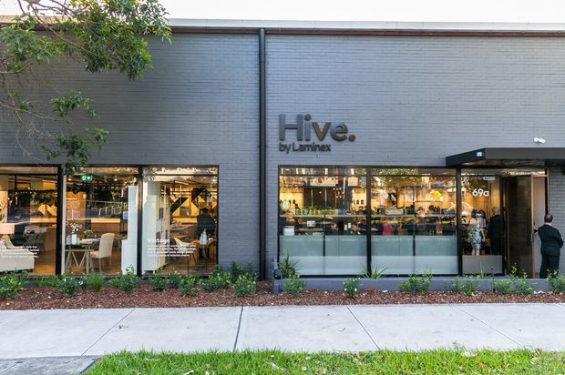 Street-facing modern commercial storefront with a dark grey textured masonry facade, large glazed display windows, and a recessed entrance beneath a projecting black canopy. The interior of the Hive showroom is visible through the glazing, with warm lighting, furniture or product displays, and signage.