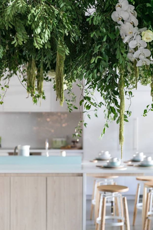 Bright contemporary kitchen and dining area with pale timber cabinetry, a light blue island benchtop, white surfaces, and timber bar stools. A large suspended arrangement of leafy greenery, trailing plants, and white orchids creates a dramatic natural focal point above the island. Soft daylight and restrained finishes give the space an airy, fresh feel.