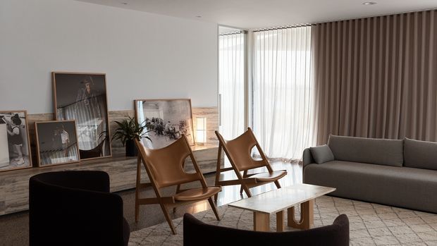 A refined contemporary living room with a grey upholstered sofa, dark lounge chairs, two leather sling chairs, and a low rectangular coffee table. Floor-to-ceiling windows are dressed with sheer curtains and full-height taupe drapes, while framed artwork rests on a low textured console along the wall. The room has a neutral, softly lit palette and a spacious, polished feel.