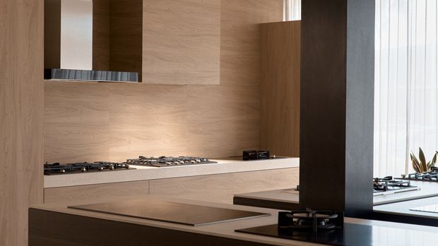 Contemporary minimalist kitchen with extensive light timber wall panelling and cabinetry, a pale stone island and benchtop, integrated gas cooktops, and a dark vertical partition framing the space. Sheer curtains and indoor plants are visible near the glazing, while a stainless-steel rangehood is partly concealed behind the cabinetry.