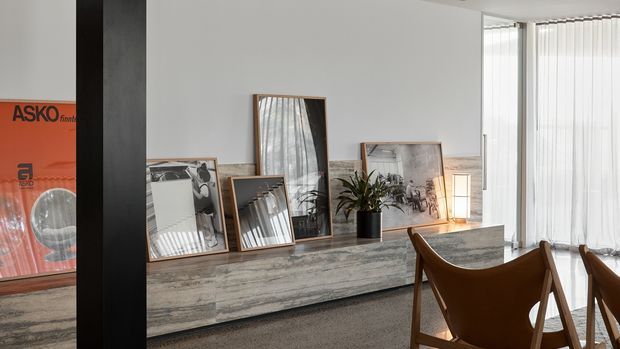 A bright contemporary interior corner featuring a long textured grey stone or concrete-look built-in console, framed artwork and photographs leaning against the wall, a potted plant, and a small table lamp. Warm timber-framed leather sling chairs occupy the foreground beside full-height windows with vertical blinds. A dark structural column divides the space, while a partially visible orange ASKO graphic appears at the far left.