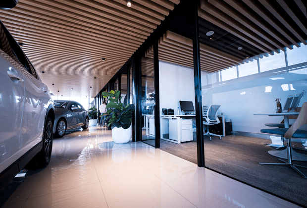 A polished contemporary commercial showroom and office interior. Vehicles are displayed along a glossy tiled circulation area on the left, while glazed private offices with desks and task chairs line the right. The space features black-framed glass partitions, a warm timber slatted ceiling, white cabinetry, indoor planting, and a bright, refined fit-out.
