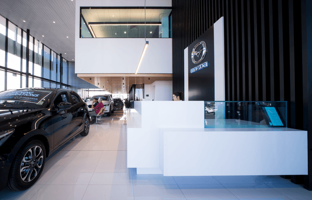 A bright, contemporary Mazda car dealership showroom with a black vehicle displayed beside a large white reception or sales counter. The space features full-height glazed windows, glossy light tile flooring, a suspended linear light, a double-height showroom volume, and a black vertical-slat feature wall with the Mazda logo. A glazed upper-level office or mezzanine is visible at the rear.