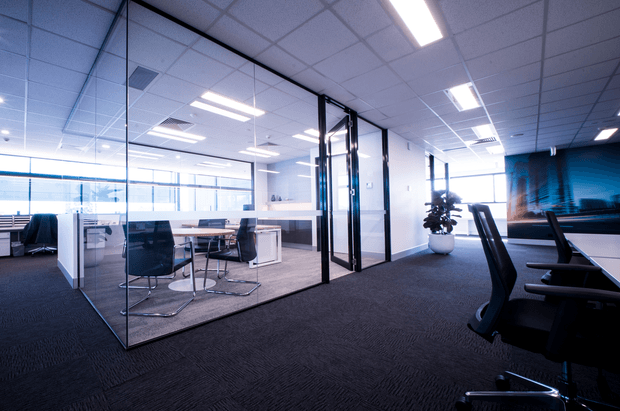 A modern commercial office interior with a carpeted open-plan workspace, glass-partitioned meeting room, desks, office chairs, bright linear ceiling lights, and large windows. The space has a cool blue and neutral palette with a clean, professional atmosphere.