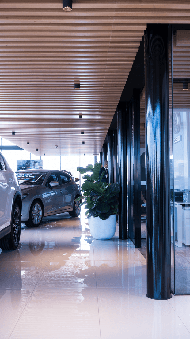 A polished automotive showroom or dealership interior with cars displayed beneath a linear timber slat ceiling. Glossy white tiled flooring reflects the ceiling and vehicles, while a row of glossy dark cylindrical columns frames glazed offices or display areas along one side. Large windows provide bright daylight, and a large potted plant adds greenery near the circulation path.