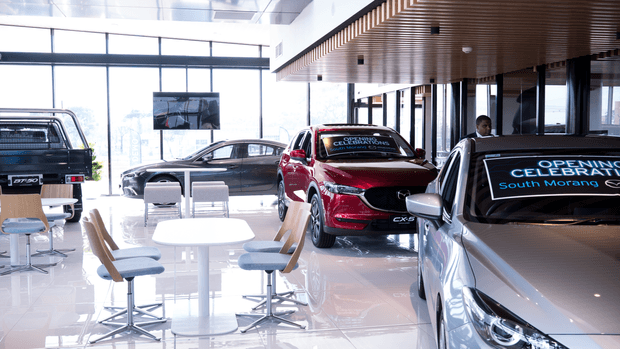 Bright contemporary automotive showroom with several display vehicles arranged around a small customer seating area. The space has floor-to-ceiling glazing, a highly polished light floor, white walls, and a distinctive slatted timber ceiling feature. Promotional lettering is visible on the vehicle windows, and a reception or sales environment is suggested.