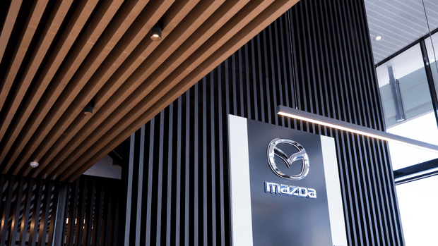 Contemporary commercial showroom or dealership interior featuring a prominent Mazda logo mounted on a dark vertically slatted feature wall. The ceiling has exposed warm timber battens with recessed spotlights, while bright linear pendant lighting and extensive glazing create a polished, high-contrast reception or display environment.