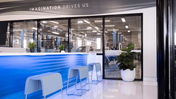 A bright contemporary commercial reception or café-style breakout area with a long blue-and-white counter, several pale blue stools, glossy white flooring, and a large potted plant. Black-framed glazed partitions reveal an industrial-style open workspace with exposed services and work areas beyond. The curved blue counter features a water-inspired graphic, and the wall above the glazing carries the text “IMAGINATION DRIVES US.”