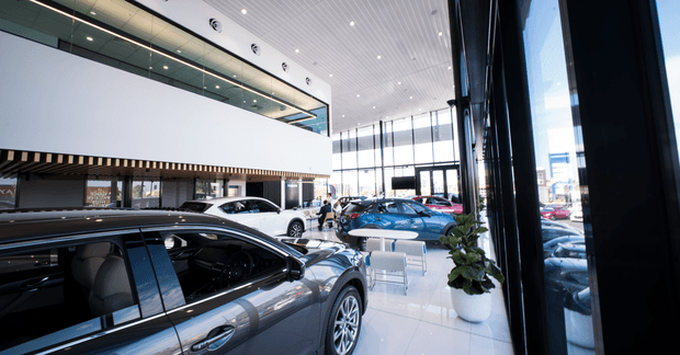 A bright, spacious modern automotive showroom viewed from inside, with several cars displayed on a glossy white floor. The space features extensive floor-to-ceiling glazing, black structural columns, a high white linear-panel ceiling with recessed spotlights, and a large white mezzanine or upper-level wall with a dark slatted edge. A small seating area and potted plant are visible near the windows.