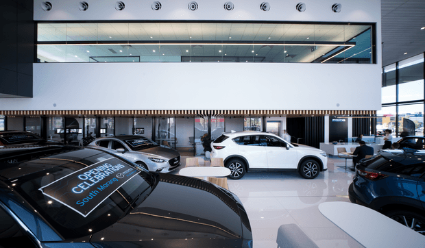 A contemporary multi-level car dealership showroom with several vehicles displayed on a glossy white tiled floor. The space features a large open volume, extensive upper-level glazing, black-framed windows, a white mezzanine-like wall, vertical timber-look screening, recessed lighting, and modern automotive display areas.