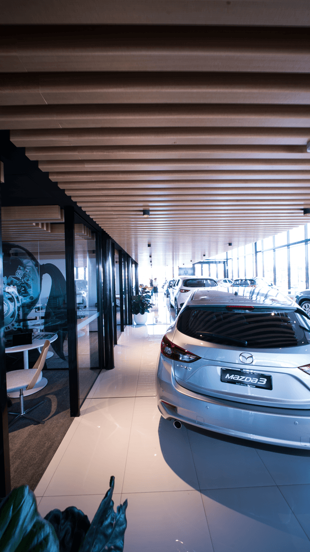 A bright contemporary automotive showroom with multiple Mazda vehicles displayed on a glossy tiled floor. The space features extensive floor-to-ceiling glazing along one side, black-framed glass office or consultation rooms, and a distinctive ceiling of closely spaced exposed timber slats. A small seating area is visible behind the glass partition.