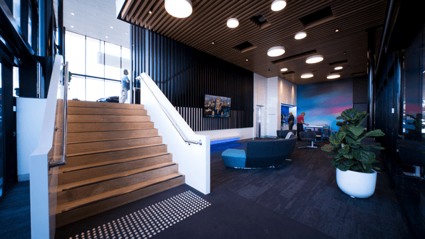 A contemporary commercial interior showing a spacious reception or lounge area beside a broad timber staircase. The space features dark carpet flooring, a slatted timber ceiling with round recessed and pendant-style lights, black vertical wall panelling, extensive floor-to-ceiling glazing, sculptural blue seating, a white planter, and a vivid blue feature wall or artwork at the rear. The atmosphere is polished, modern, and professional.