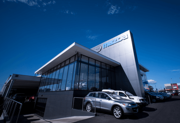 Exterior architectural photograph of a modern Mazda automotive dealership or showroom. The building features a sharply angled silver metal-clad facade with prominent Mazda signage, extensive floor-to-ceiling dark-framed glazing, a broad projecting flat roof, and a paved forecourt displaying vehicles.