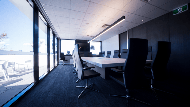A contemporary commercial meeting room with a long white conference table surrounded by high-back black and white office chairs. Full-height glazing opens toward a bright outdoor terrace, while a dark feature wall, linear pendant lighting, carpeted floor, and suspended acoustic tile ceiling create a sleek corporate interior.