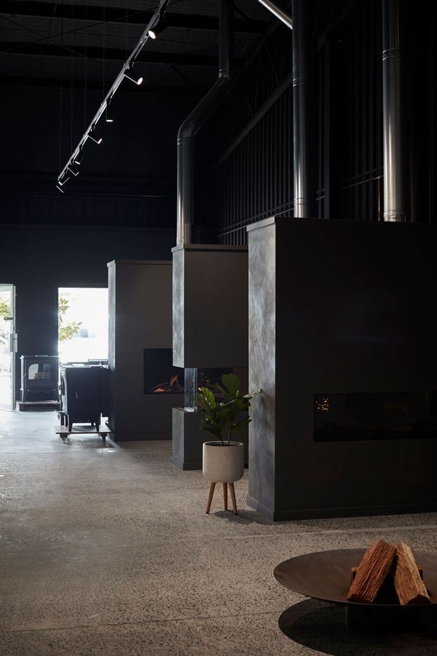 A moody industrial-style interior with dark metal walls, exposed ducts and ceiling structure, track lighting, polished concrete flooring, and multiple sculptural built-in fireplaces. A potted plant, lounge chair, and low firewood table add warmth to the dramatic space.