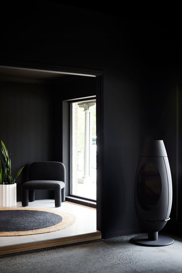 A dark, moody contemporary interior featuring a black-paneled room opening into a brighter sitting area. The space includes a sculptural black freestanding fireplace, a rounded charcoal lounge chair, a tall indoor plant in a white planter, a circular two-tone rug, dark-framed glazing, and a light tiled floor in the adjoining area.
