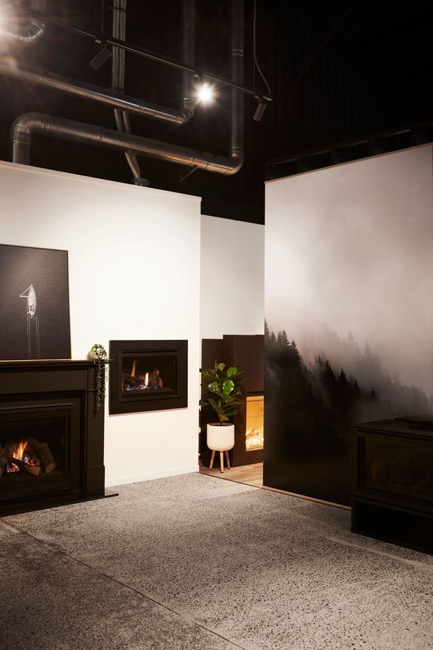 A dark, dramatic showroom or interior space featuring multiple built-in fireplaces set against white walls, black cabinetry, exposed metallic ductwork, track lighting, and a large misty forest mural. The floor has a speckled polished concrete or terrazzo-like finish, with a potted plant and warm firelight adding contrast.