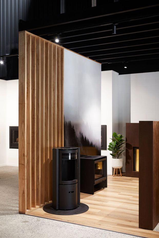A contemporary showroom interior displaying freestanding heating appliances and fireplaces. The space features a tall vertical timber-slat partition, a large white graphic feature panel, black exposed ceiling structure with track lighting, pale timber flooring, white walls, and several dark fireplaces and wood-burning heaters. A potted plant and additional fireplace displays are visible in the background.