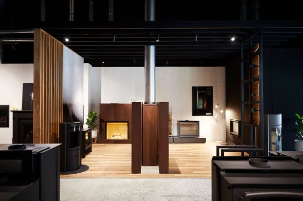 A dark, sophisticated contemporary interior with a split-level open-plan living and display space. Black exposed ceiling beams and structural framing contrast with white walls, warm timber flooring, vertical timber screening, dark cabinetry, and multiple fireplace or heating displays. The room includes built-in joinery, shelving, lighting, and a central timber partition.